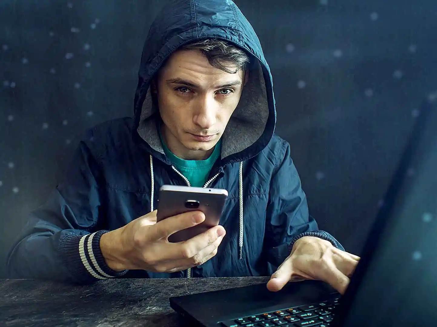 A person in a hoodie holding a phone and using a computer